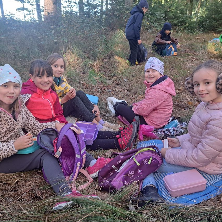 Fünf junge Mädchen sitzen auf dem Boden in einem Wald. Sie tragen Winterkleidung und haben Rucksäcke. Einige lächeln. Hinter ihnen stehen andere Leute, eine davon steht.