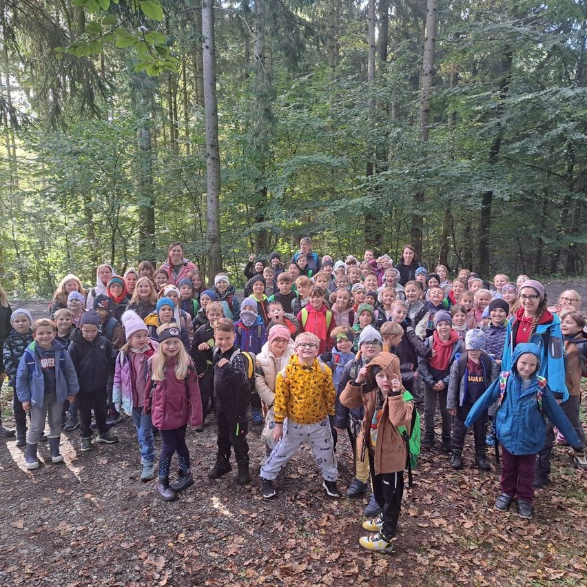 Eine Gruppe von Kindern und Erwachsenen steht in einem Waldgebiet, um ein Foto zu machen, mit einem Lehmweg im Vordergrund und Bäumen dahinter.