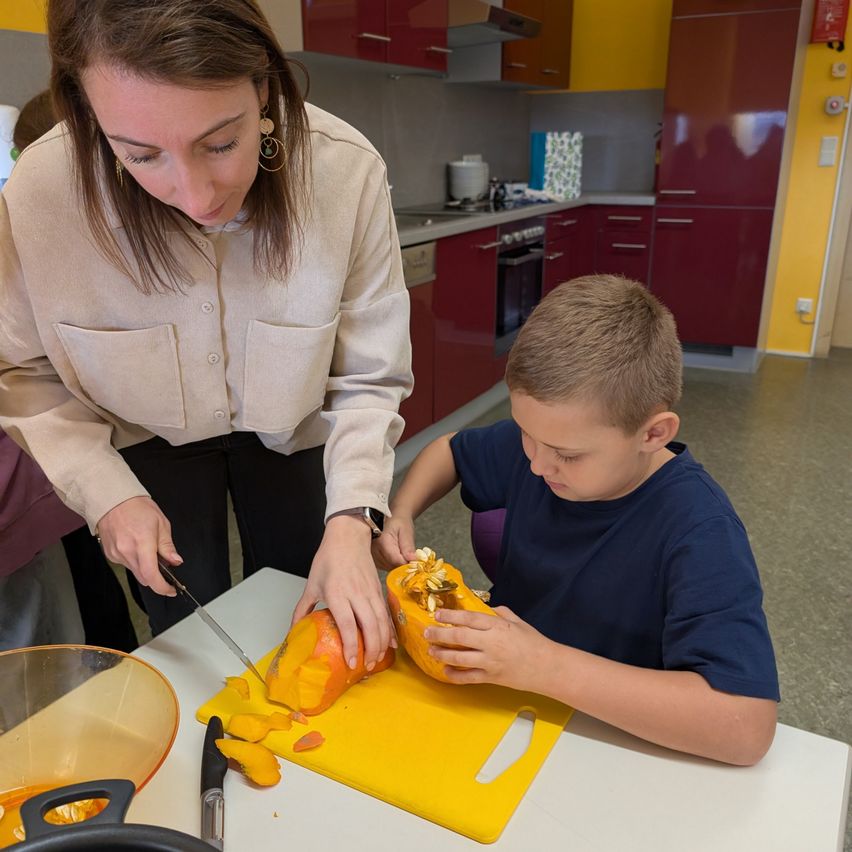 Eine Frau schneidet eine Kürbis, während ein Junge den Kürbis auf einem gelben Schneidebrett auf einem Tisch hält.