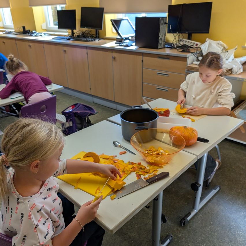 Drei Mädchen sind in einem Klassenzimmer, jedes schneidet Kürbisse auf seinen Tischen. Die Tische sind weiß mit Schneidebrettern, Messern und Schüsseln. Das Klassenzimmer hat Schränke, Monitore und Stühle.