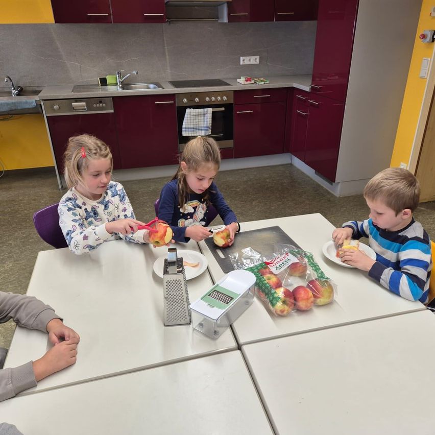 Drei Kinder sitzen um einen Tisch in einer Küche und schälen Äpfel. Ein Kind benutzt einen Schäler, während ein anderes ein Messer benutzt. Eine Tüte Äpfel steht auf dem Tisch.