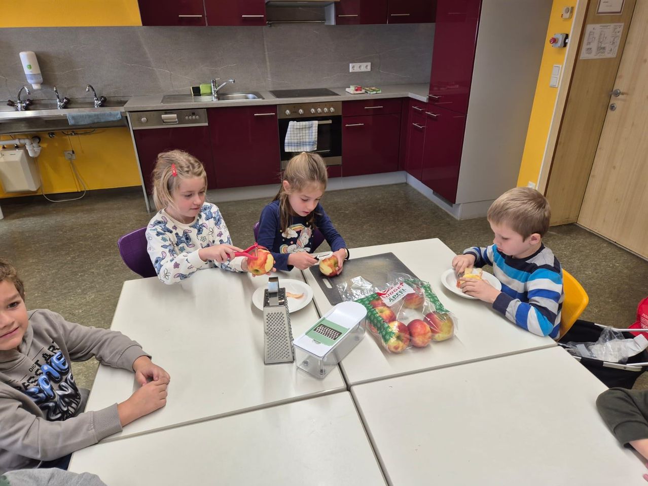 Drei Kinder sitzen um einen Tisch in einer Küche und schälen Äpfel. Ein Kind benutzt einen Schäler, während ein anderes ein Messer benutzt. Eine Tüte Äpfel steht auf dem Tisch.