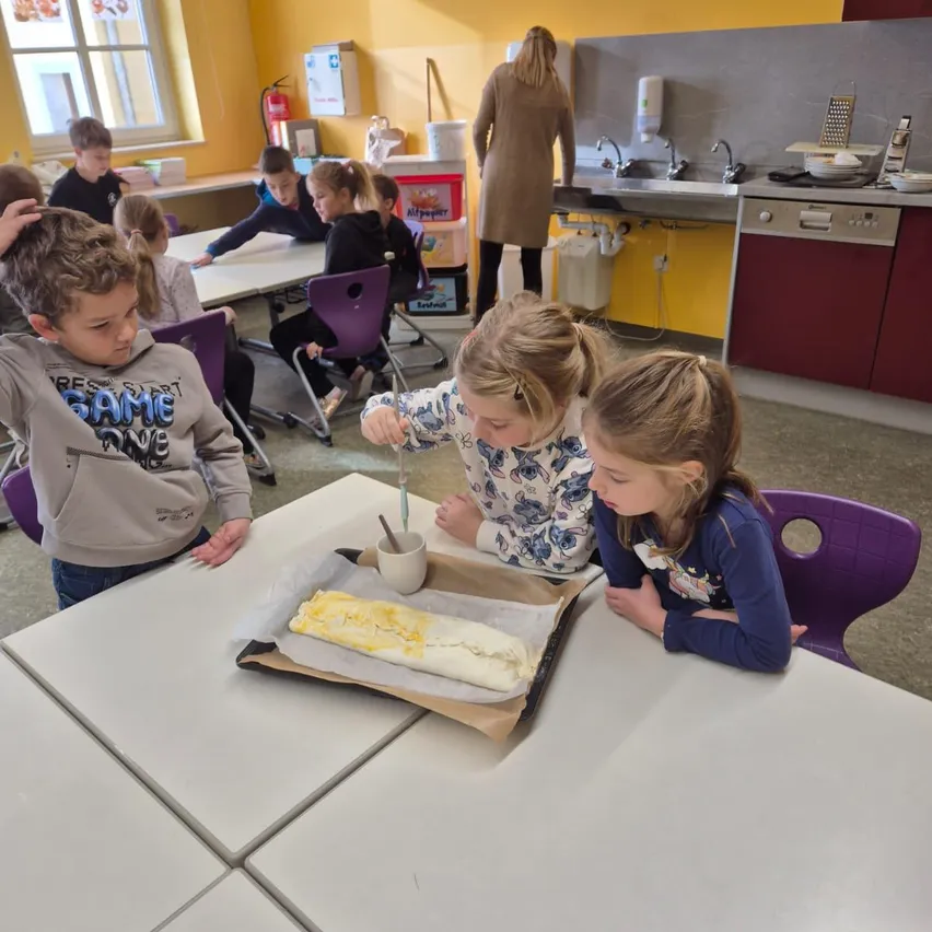 Kinder in einem Klassenzimmer arbeiten an einem Backprojekt. Zwei Mädchen bestreichen ein Gebäck, während ein Junge zuschaut. Andere Kinder sitzen im Hintergrund an Tischen.
