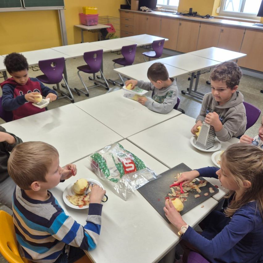 Eine Gruppe von Kindern in einem Klassenzimmer sitzt an Tischen und isst Äpfel. Ein Mädchen schneidet einen Apfel mit einem Schäler auf einem Schneidebrett.