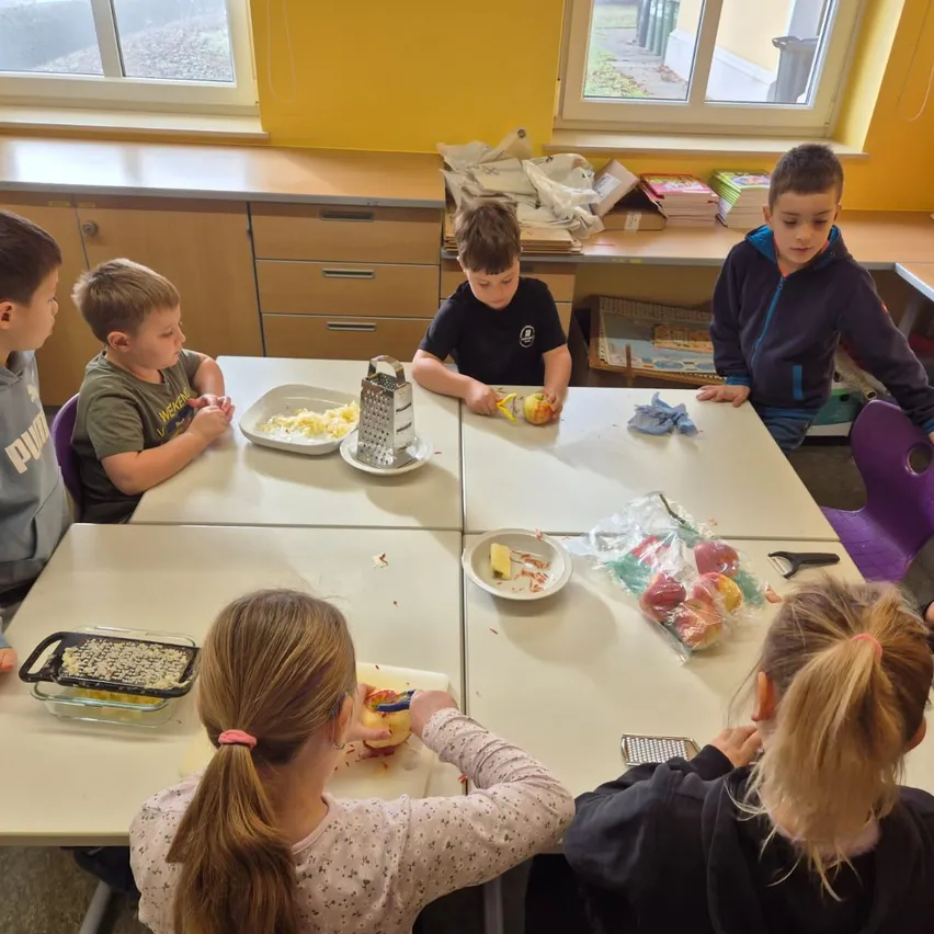 Kinder in einem Klassenzimmer, die um einen Tisch herum sitzen, schälen Äpfel. Ein Kind hält einen Apfel und einen Schäler. Hinter ihnen sind Regale mit Büchern und Kisten zu sehen.