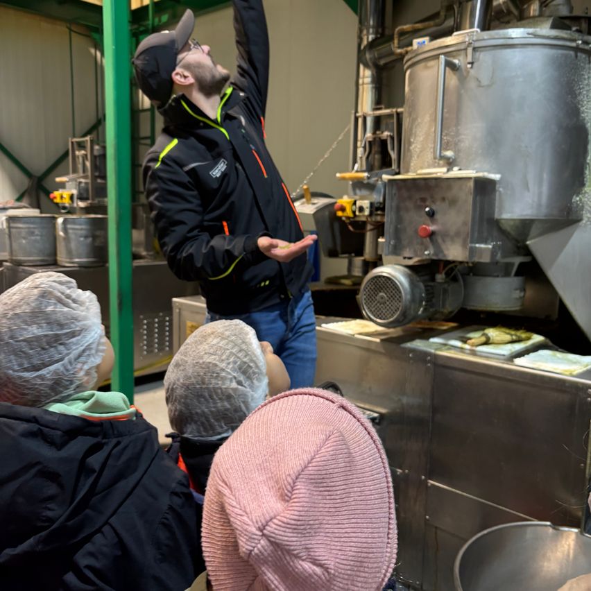 Ein Mann in einer schwarzen Jacke zeigt auf eine Maschine, während Kinder dahinter zusehen.