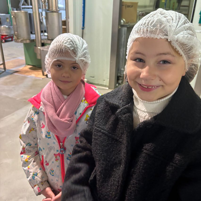 Zwei Mädchen stehen in einer Fabrik und tragen Haarnetze. Das Mädchen auf der linken Seite trägt einen rosa Schal und eine bedruckte Jacke, während das Mädchen auf der rechten Seite einen schwarzen Mantel trägt. Hinter ihnen steht eine Metallkonstruktion mit einem Behälter.
