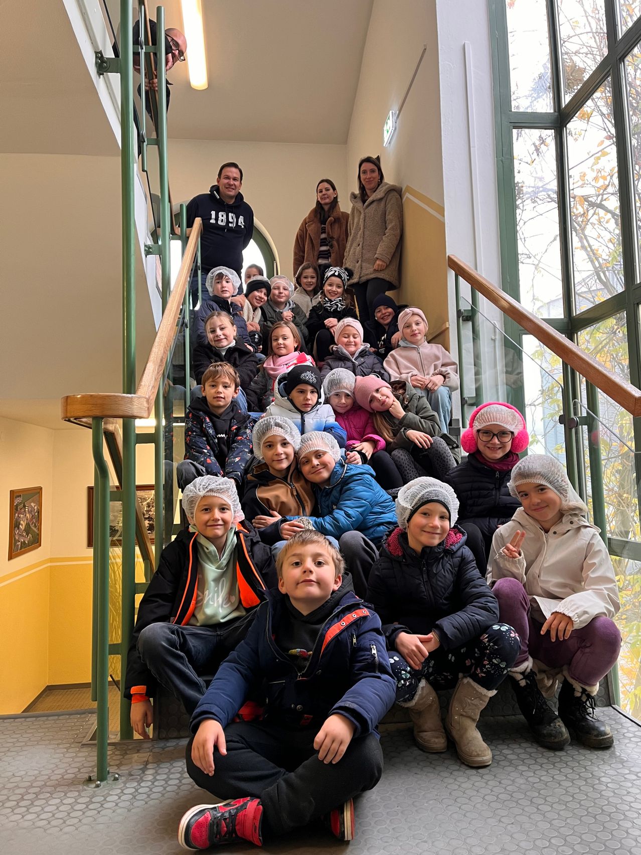 Eine Gruppe von Kindern in Winterkleidung und Haarnetzen sitzt auf einer Treppe. Zwei Erwachsene stehen oben an der Treppe, mit weiteren Kindern darunter. Die Umgebung umfasst Glasfenster und ein hölzerne Handlauf.