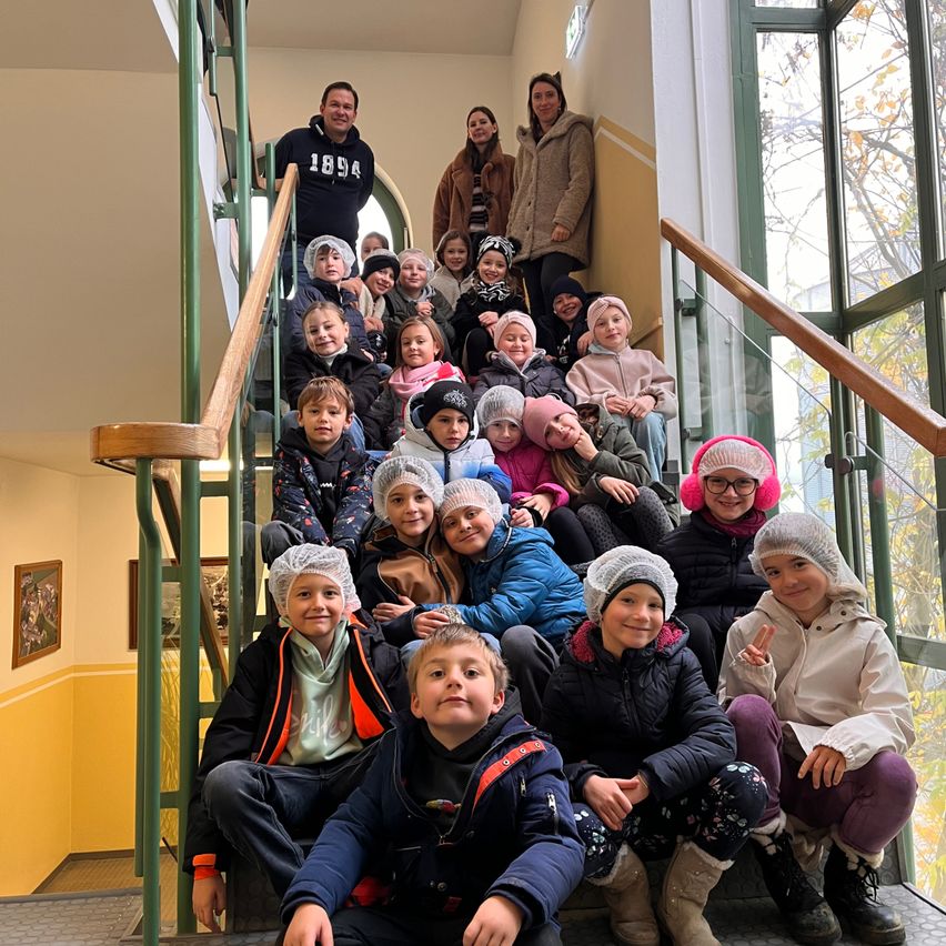 Eine Gruppe von Kindern in Winterkleidung und Haarnetzen sitzt auf einer Treppe. Zwei Erwachsene stehen oben an der Treppe, mit weiteren Kindern darunter. Die Umgebung umfasst Glasfenster und ein hölzerne Handlauf.