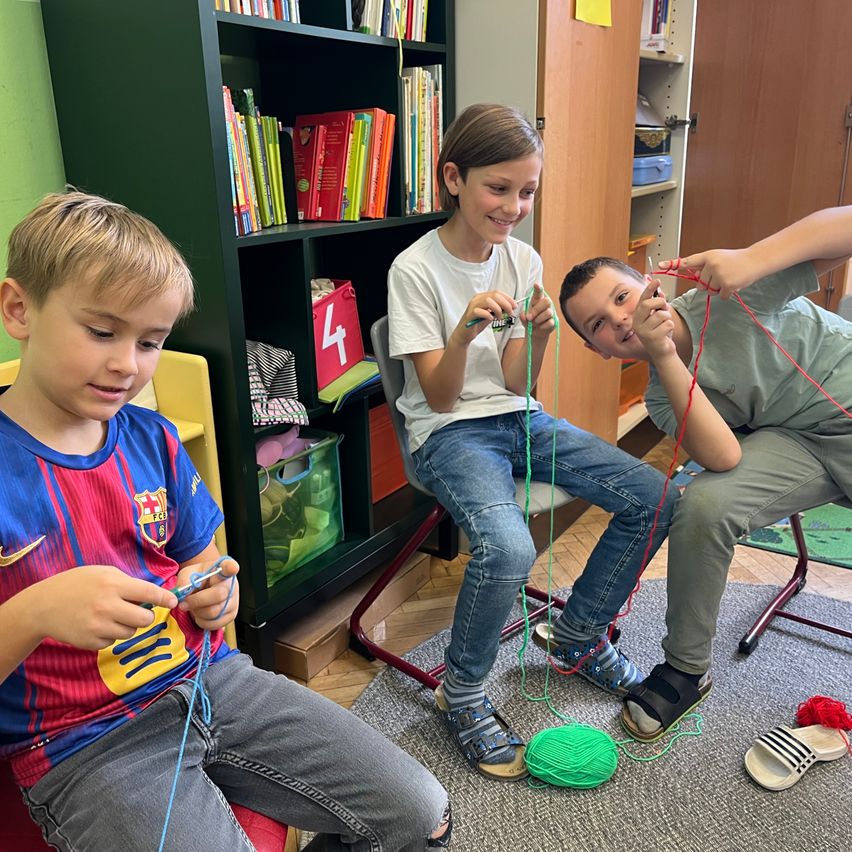 Drei Jungen sitzen in einem Raum und stricken mit Garn. Der Junge auf der linken Seite trägt ein Barcelona-Fußballtrikot. Dahinter steht ein Bücherregal voller Bücher.