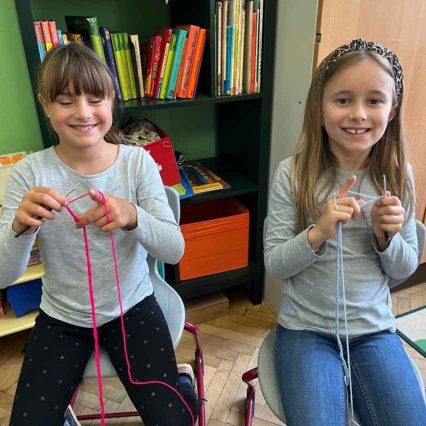 Zwei junge Mädchen sitzen auf Stühlen und stricken mit Garn. Das Mädchen auf der linken Seite hält ein rosa Garn und das Mädchen auf der rechten Seite hält ein blaues Garn. Hinter ihnen steht ein Bücherregal mit vielen Büchern.