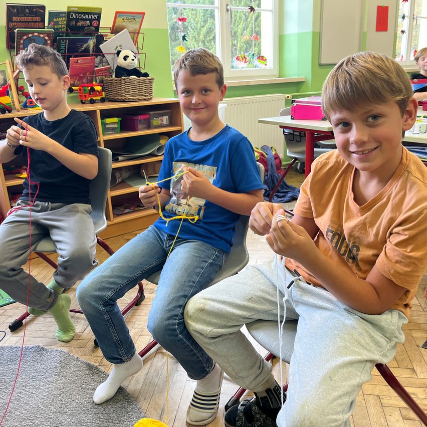 Drei Jungen sitzen in Stühlen in einem Klassenzimmer und arbeiten an einem Fadenprojekt. Der Junge auf der linken Seite hält einen roten Faden, der in der Mitte einen gelben Faden und der Junge auf der rechten Seite einen weißen Faden. Sie tragen Freizeitkleidung. Hinter ihnen sind Regale mit Büchern und Spielzeug zu sehen.