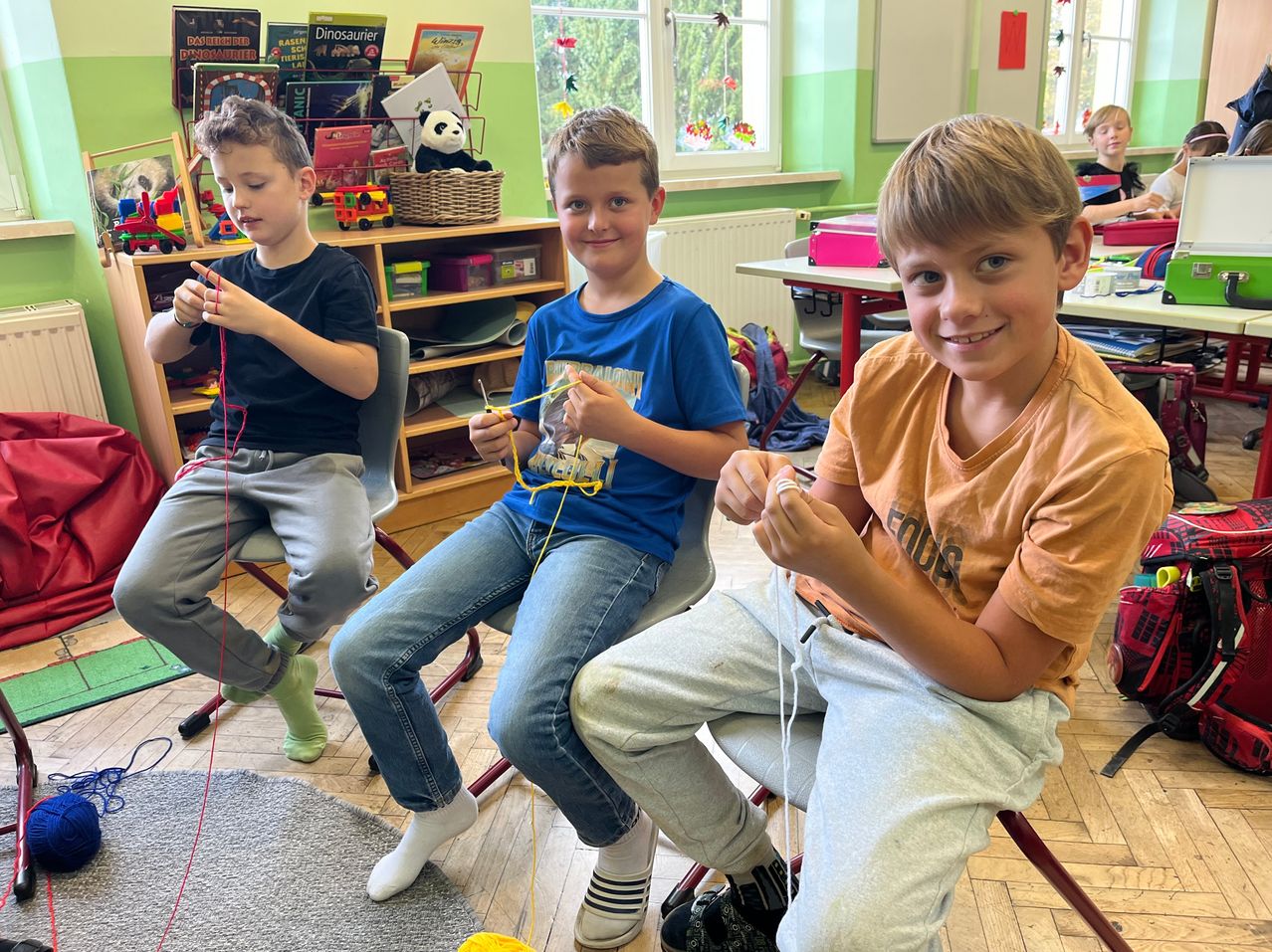 Drei Jungen sitzen in Stühlen in einem Klassenzimmer und arbeiten an einem Fadenprojekt. Der Junge auf der linken Seite hält einen roten Faden, der in der Mitte einen gelben Faden und der Junge auf der rechten Seite einen weißen Faden. Sie tragen Freizeitkleidung. Hinter ihnen sind Regale mit Büchern und Spielzeug zu sehen.