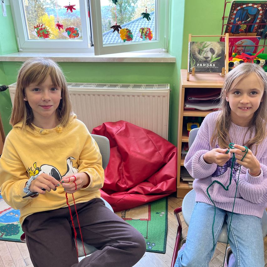 Zwei junge Mädchen sitzen in Stühlen und stricken mit rotem und grünem Garn. Hinter ihnen ist ein Fenster mit bunten Herbstdekorationen. Ein Buch über Pandas steht auf einem Regal, und ein roter Sitzsack ist in der Nähe.