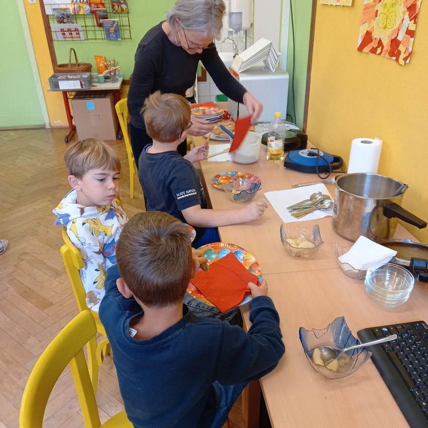 Drei Jungen sitzen an einem Tisch und bereiten Essen zu. Eine ältere Frau hilft und hält ein rotes Papier. Schüsseln, Teller und Besteck sind auf dem Tisch, zusammen mit einem Topf und einer Rolle Küchenpapier.