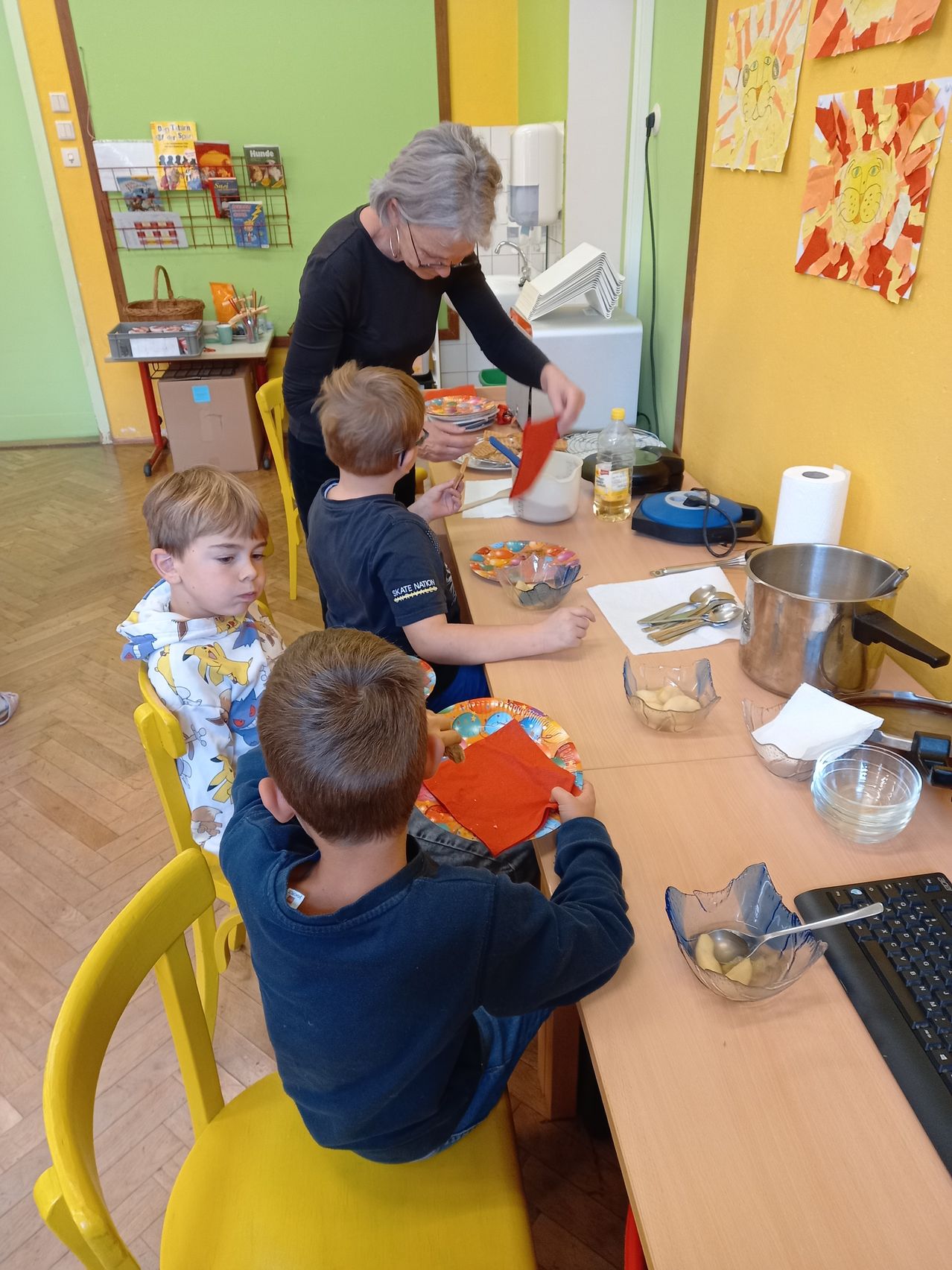Drei Jungen sitzen an einem Tisch und bereiten Essen zu. Eine ältere Frau hilft und hält ein rotes Papier. Schüsseln, Teller und Besteck sind auf dem Tisch, zusammen mit einem Topf und einer Rolle Küchenpapier.