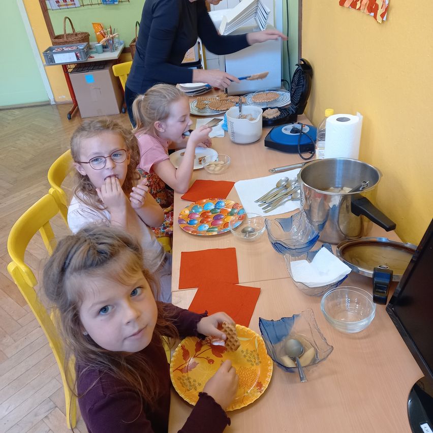 Drei junge Mädchen sitzen an einem Tisch und essen. Eine Frau steht hinter dem Tisch und scheint ihnen zu servieren. Der Tisch hat Teller, Schüsseln, Besteck und einen Monitor darauf.