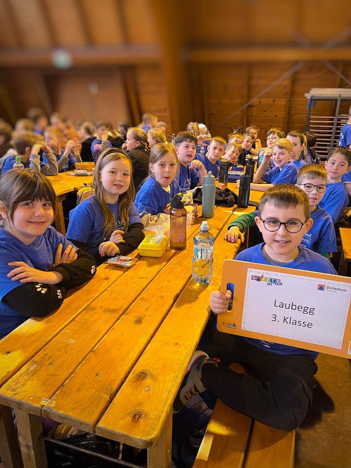 Schüler sitzen an einem langen Holztisch in einem Essbereich. Ein Junge hält ein Schild mit der Aufschrift 'Lauegg 3. Klasse.' Flaschen und eine Lunchbox stehen auf dem Tisch.