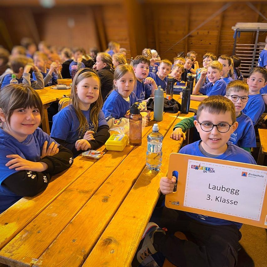 Schüler sitzen an einem langen Holztisch in einem Essbereich. Ein Junge hält ein Schild mit der Aufschrift 'Lauegg 3. Klasse.' Flaschen und eine Lunchbox stehen auf dem Tisch.