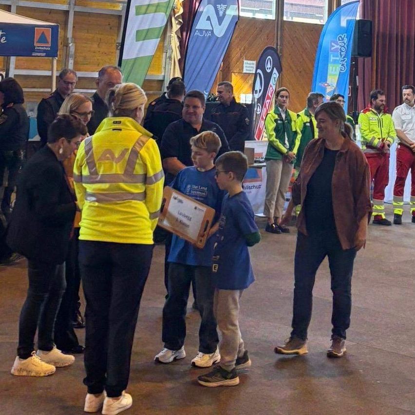 Eine Gruppe von Menschen steht in einem Raum mit zwei Kindern, die eine Plakette halten. Eine Frau in einer gelben Sicherheitsjacke überreicht die Plakette an die Kinder. Hinter ihnen stehen mehrere Personen in Sicherheitsjacken und Uniformen.