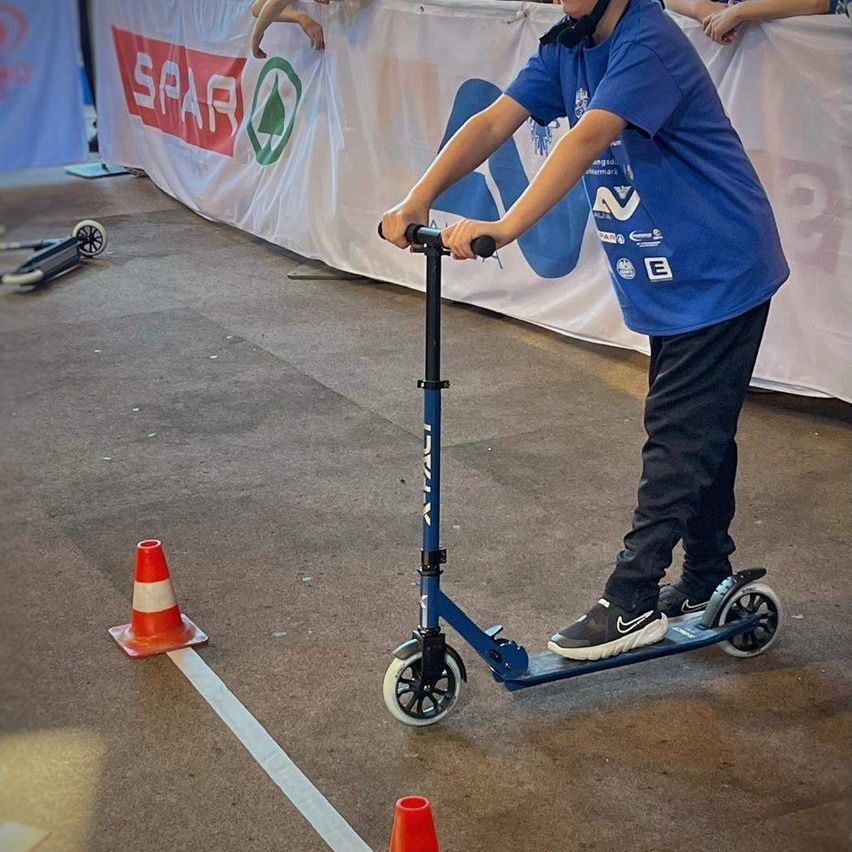 Ein junger Junge fährt einen blauen Roller auf einem Betonboden und passiert einen orangefarbenen Verkehrskegel. Ein weißes Banner mit dem Wort SPAR ist im Hintergrund.