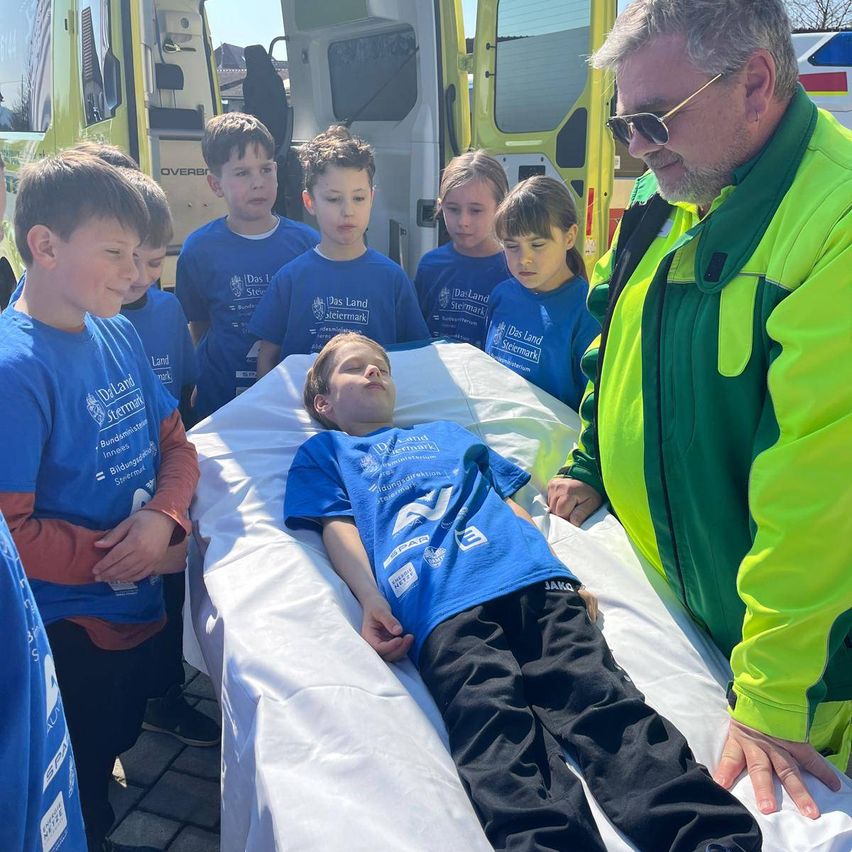 Ein Junge liegt auf einer Trage, daneben ein Krankenwagenpersonal. Mehrere Kinder in blauen Shirts stehen herum, eines hält ein Schild. Der Mann im grünen Jackett schaut aufmerksam zu.