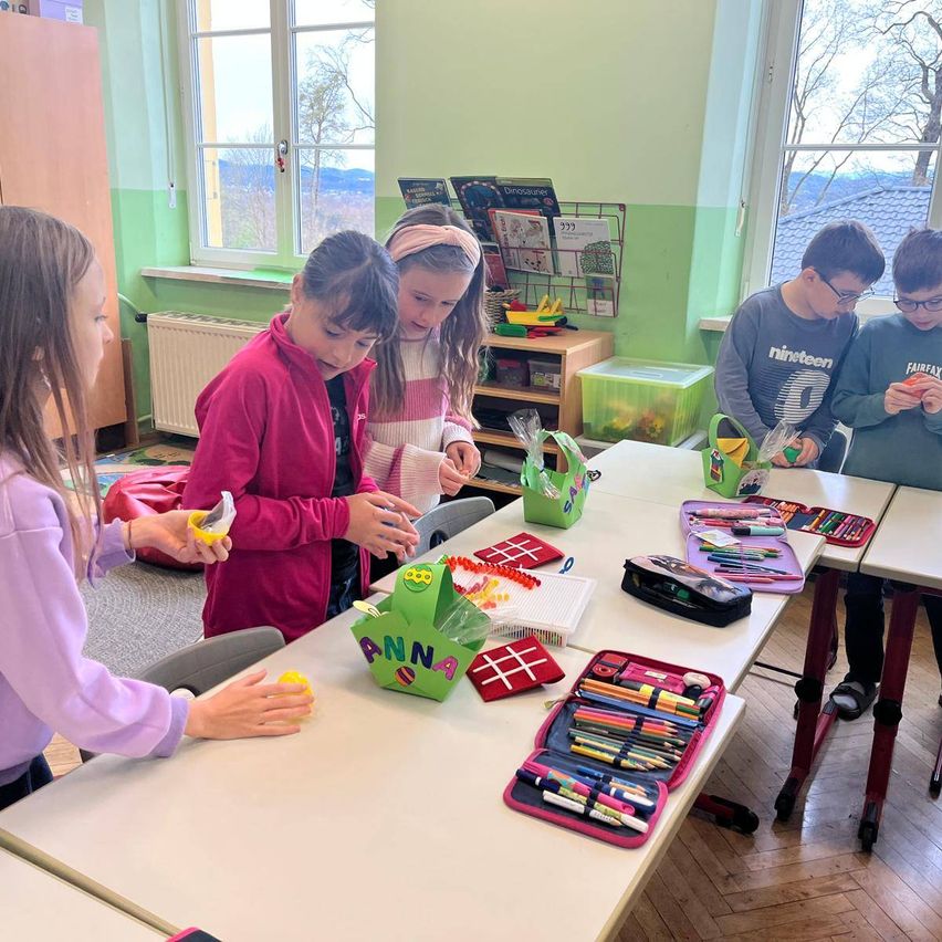 Fünf Kinder in einem Klassenzimmer, drei Mädchen dekorieren einen Osterkorb, während zwei Jungen etwas betrachten. Es gibt Kunstmaterialien auf dem Tisch.