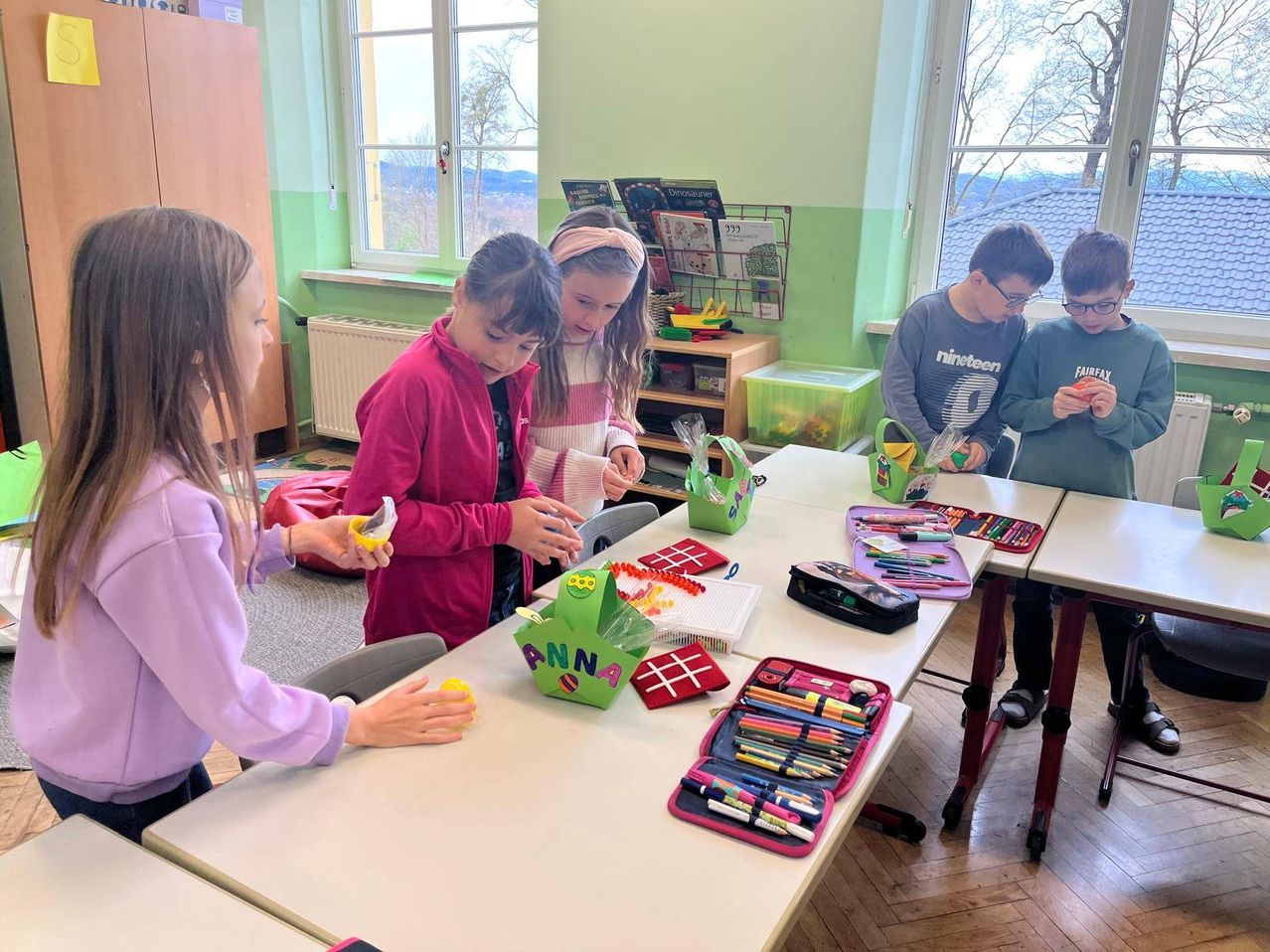 Fünf Kinder in einem Klassenzimmer, drei Mädchen dekorieren einen Osterkorb, während zwei Jungen etwas betrachten. Es gibt Kunstmaterialien auf dem Tisch.