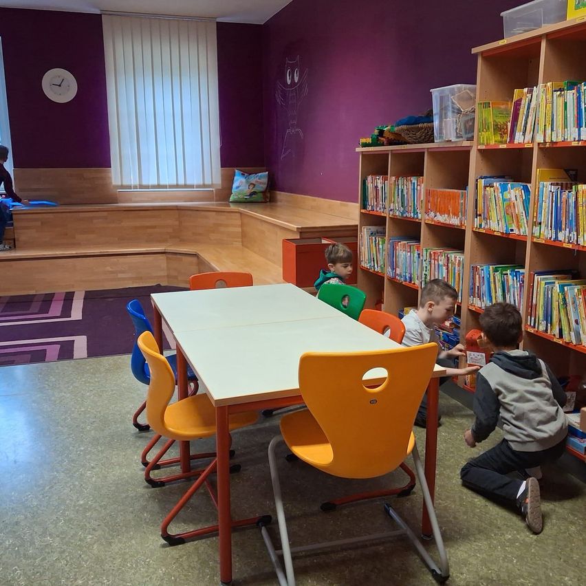 Vier Kinder sitzen in einem Raum mit einem weißen Tisch und orangefarbenen Stühlen. Ein Junge kniet vor einem Regal mit Büchern.