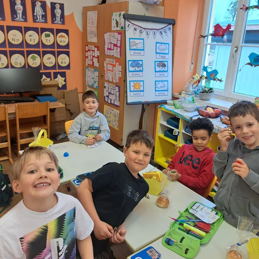 Fünf Jungen sitzen um einen Schreibtisch in einem Klassenzimmer. Ein Junge vorne hat ein farbenfrohes T-Shirt. Ein anderer Junge hält einen Plastikbeutel. Ein Whiteboard mit englischen Wörtern steht hinter ihnen.