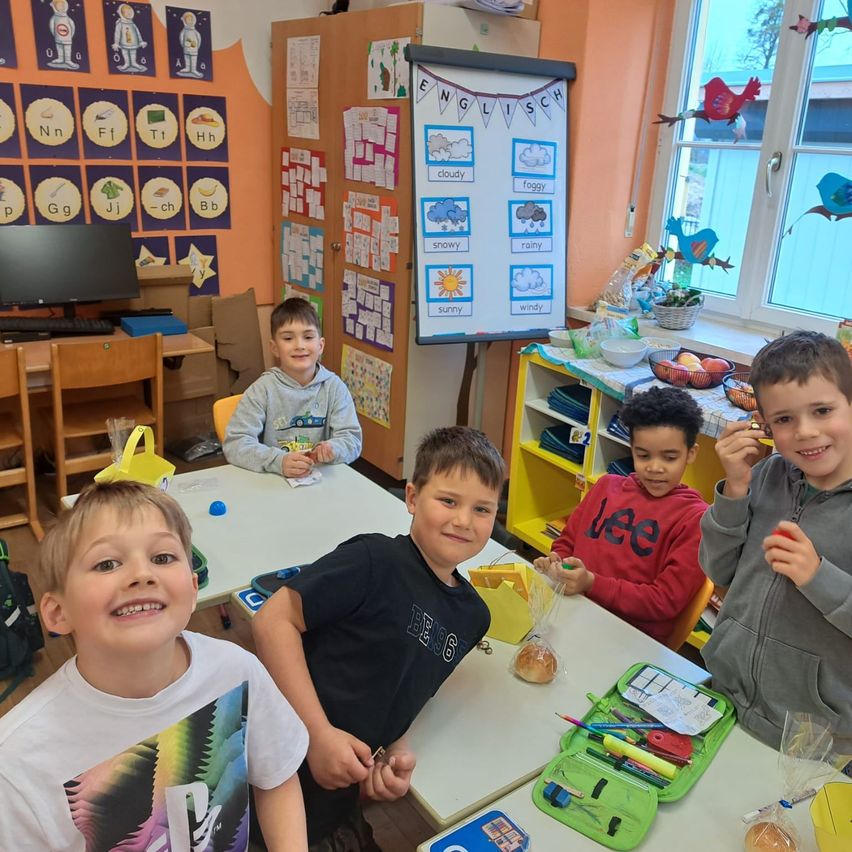 Fünf Jungen sitzen um einen Schreibtisch in einem Klassenzimmer. Ein Junge vorne hat ein farbenfrohes T-Shirt. Ein anderer Junge hält einen Plastikbeutel. Ein Whiteboard mit englischen Wörtern steht hinter ihnen.