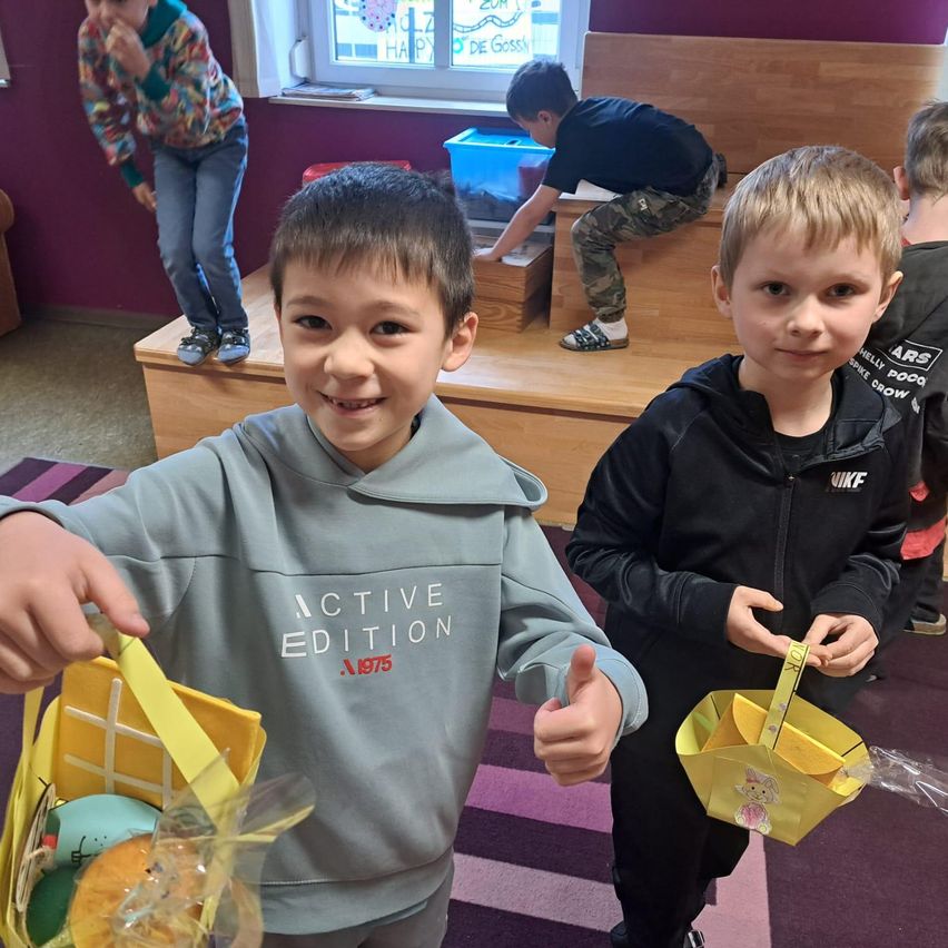 Zwei Jungen halten Osterkörbchen. Ein Junge hat eine Daumen hoch. Eine Mädchen isst im Hintergrund etwas und ein Junge klettert auf eine Holzstufe.