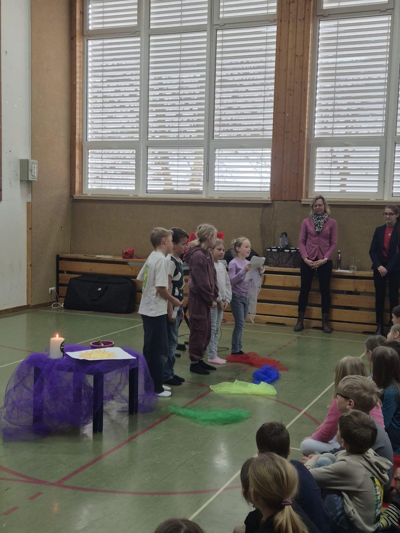 Kinder stehen um eine Kerze auf einem Tisch, während andere auf einer Bühne stehen. Eine Frau hält ein Papier und eine Handtasche. Der Raum hat Holzwände, Fenster und einen Boden mit mehrfarbigen Bändern.
