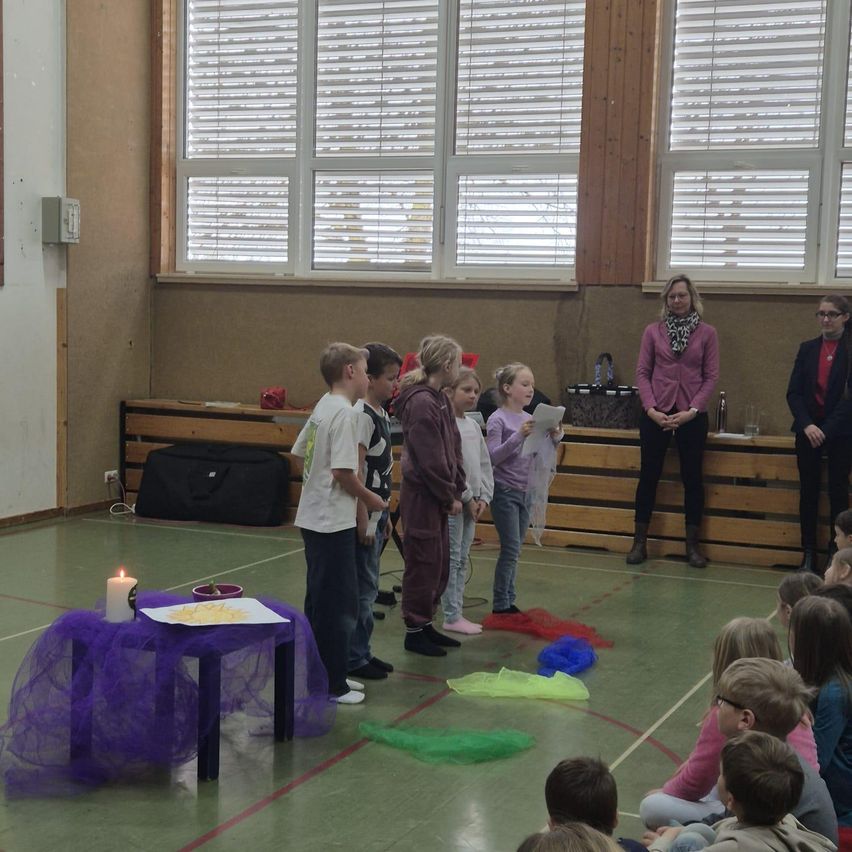 Kinder stehen um eine Kerze auf einem Tisch, während andere auf einer Bühne stehen. Eine Frau hält ein Papier und eine Handtasche. Der Raum hat Holzwände, Fenster und einen Boden mit mehrfarbigen Bändern.
