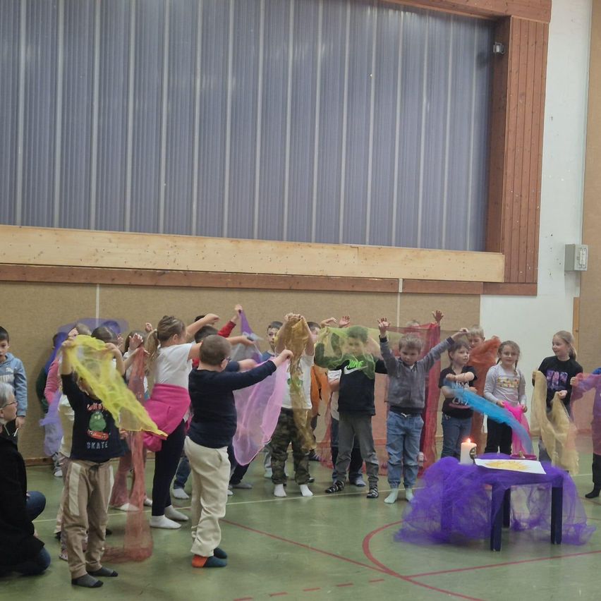 Kinder üben einen Tanz in einer Turnhalle mit bunten Tüchern in den Händen. Auf einem mit lila Stoff bedeckten Tisch steht eine Kerze.
