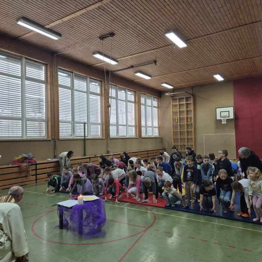 Eine Gruppe von Kindern und Erwachsenen versammelt sich in einer Turnhalle für eine Zeremonie. Ein Priester leitet die Zeremonie mit einer Kerze auf einem kleinen Tisch. Die Kinder knien auf blauen Matten.