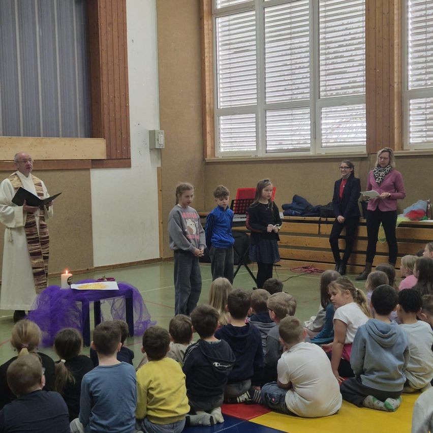 Ein Priester steht vor einer Menschenmenge von Kindern mit einer Kerze auf einem Tisch, während zwei Frauen hinter den Kindern stehen.