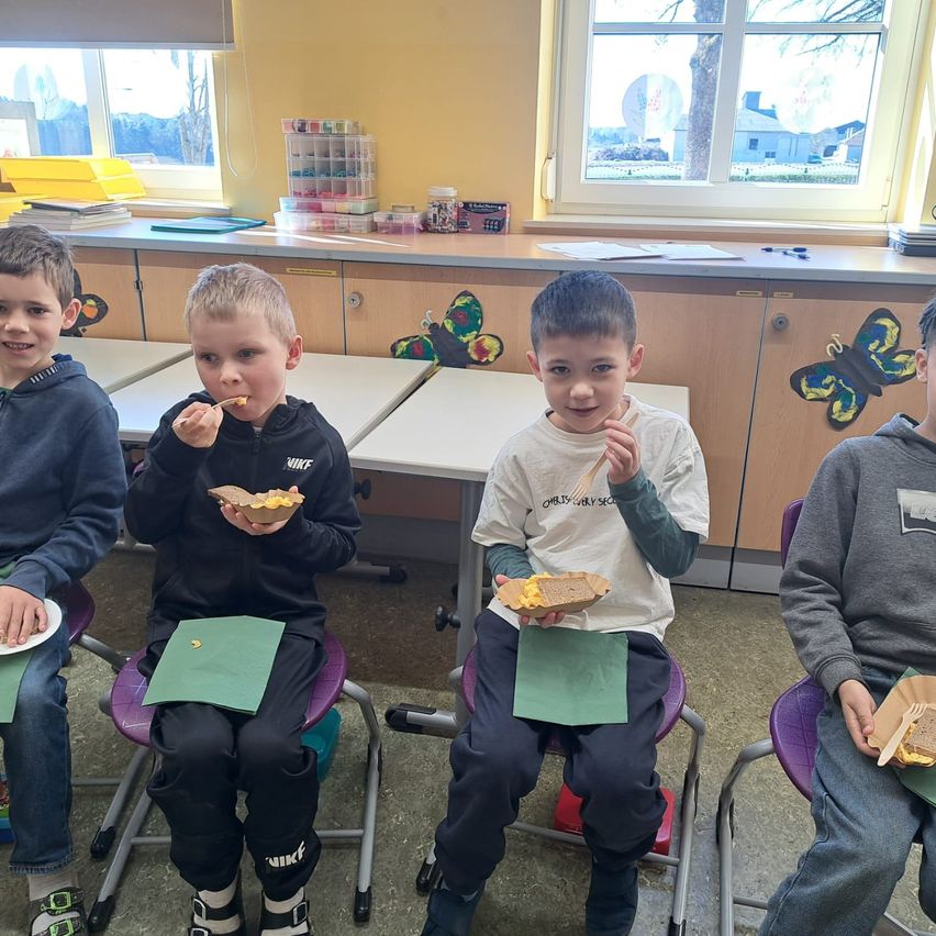 Vier Jungen sitzen in einem Klassenzimmer und essen Sandwiches auf Papiertellern. Zwei halten Gabeln, einer benutzt einen Löffel. Fenster mit Schmetterlingsausschnitten sind hinter ihnen.
