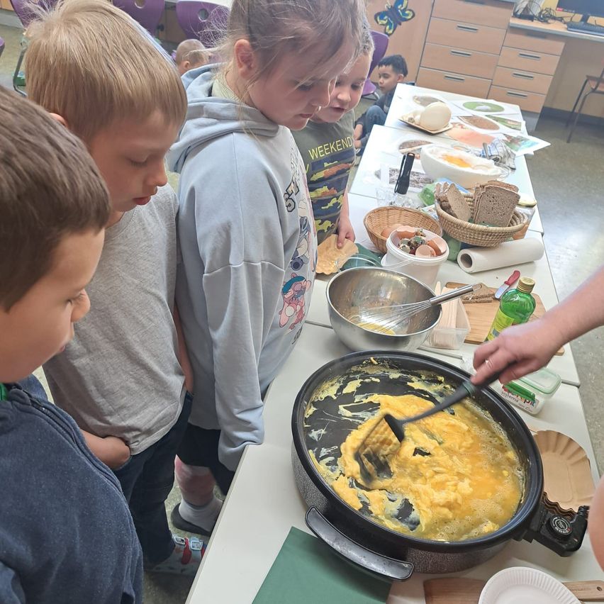 Eine Gruppe von Kindern steht um einen Tisch herum und beobachtet, wie jemand Rührei kocht. Schüsseln, Teller und Zutaten liegen auf dem Tisch.