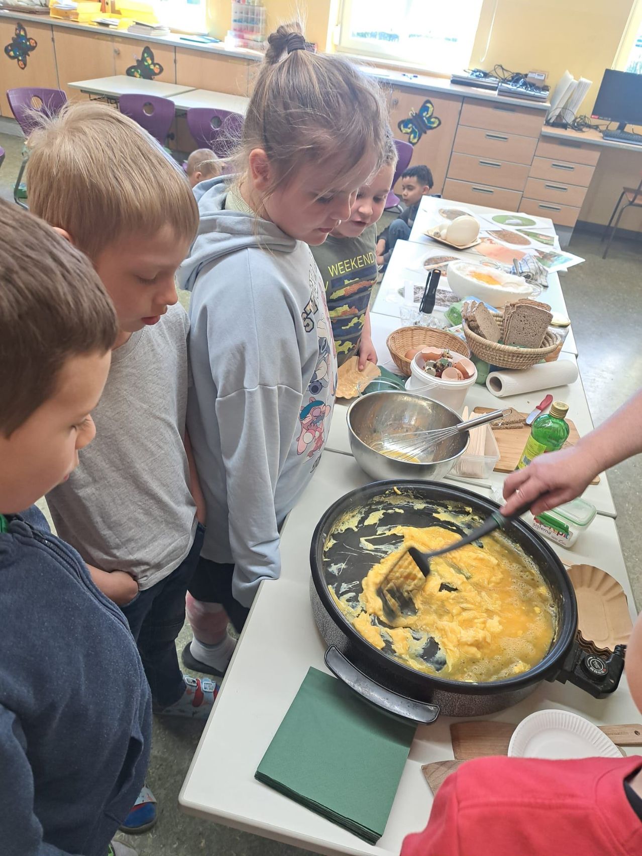 Eine Gruppe von Kindern steht um einen Tisch herum und beobachtet, wie jemand Rührei kocht. Schüsseln, Teller und Zutaten liegen auf dem Tisch.