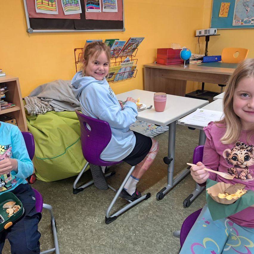 Drei Kinder sitzen an einem Tisch im Klassenzimmer. Das Mädchen in der blauen Kapuzenjacke lächelt, während die anderen beiden Kinder Essen in den Händen haben. Ein Globus und ein Projektor stehen auf dem Lehrertisch.