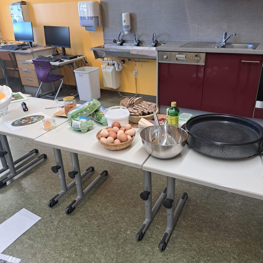 Eine Küchenecke mit aufgestellten Klapptischen, auf denen verschiedene Zutaten bereitstehen. Es gibt Eier, Brot und Gemüse auf dem Tisch, zusammen mit Küchenutensilien wie einer Bratpfanne und einer Schüssel.