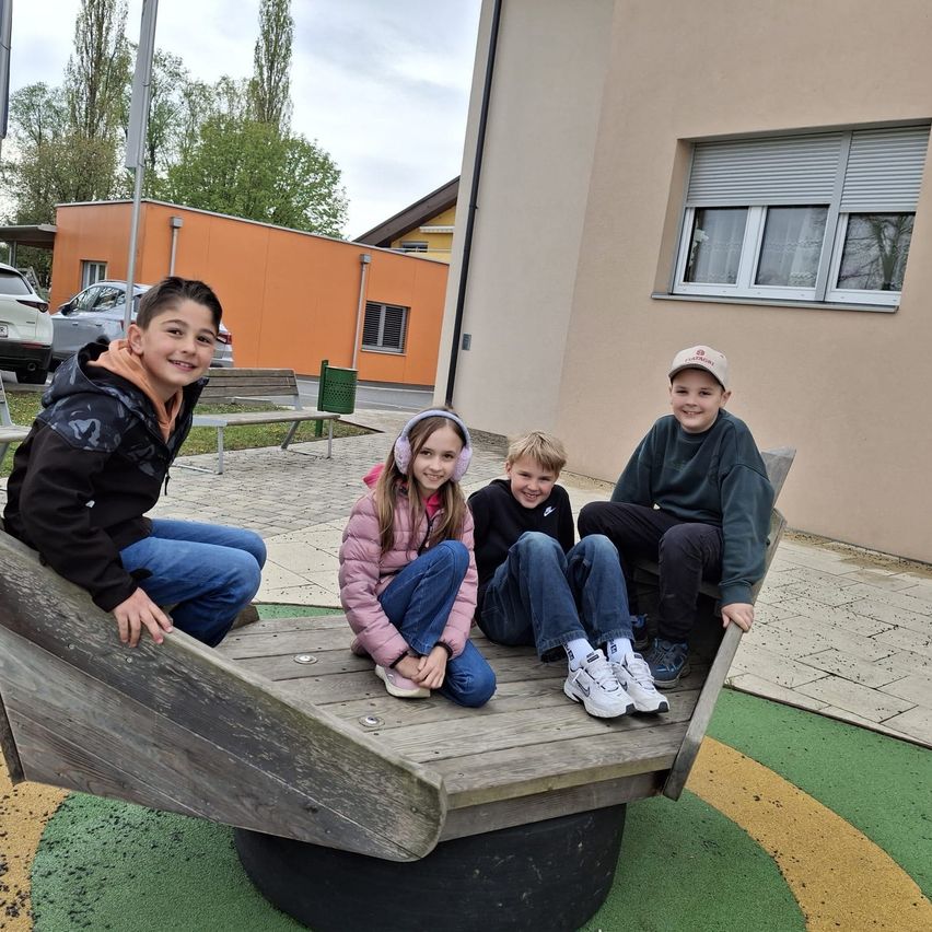 Vier Kinder sitzen auf einer kreisförmigen Attraktion im Spielplatz. Ein Junge trägt einen Hut, ein anderer Kopfhörer. Ein Mädchen trägt eine rosa Jacke. Hinter ihnen hat ein Gebäude ein Fenster mit Jalousien.