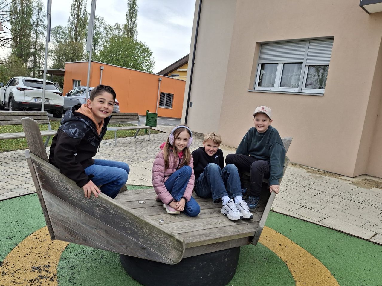 Vier Kinder sitzen auf einer kreisförmigen Attraktion im Spielplatz. Ein Junge trägt einen Hut, ein anderer Kopfhörer. Ein Mädchen trägt eine rosa Jacke. Hinter ihnen hat ein Gebäude ein Fenster mit Jalousien.