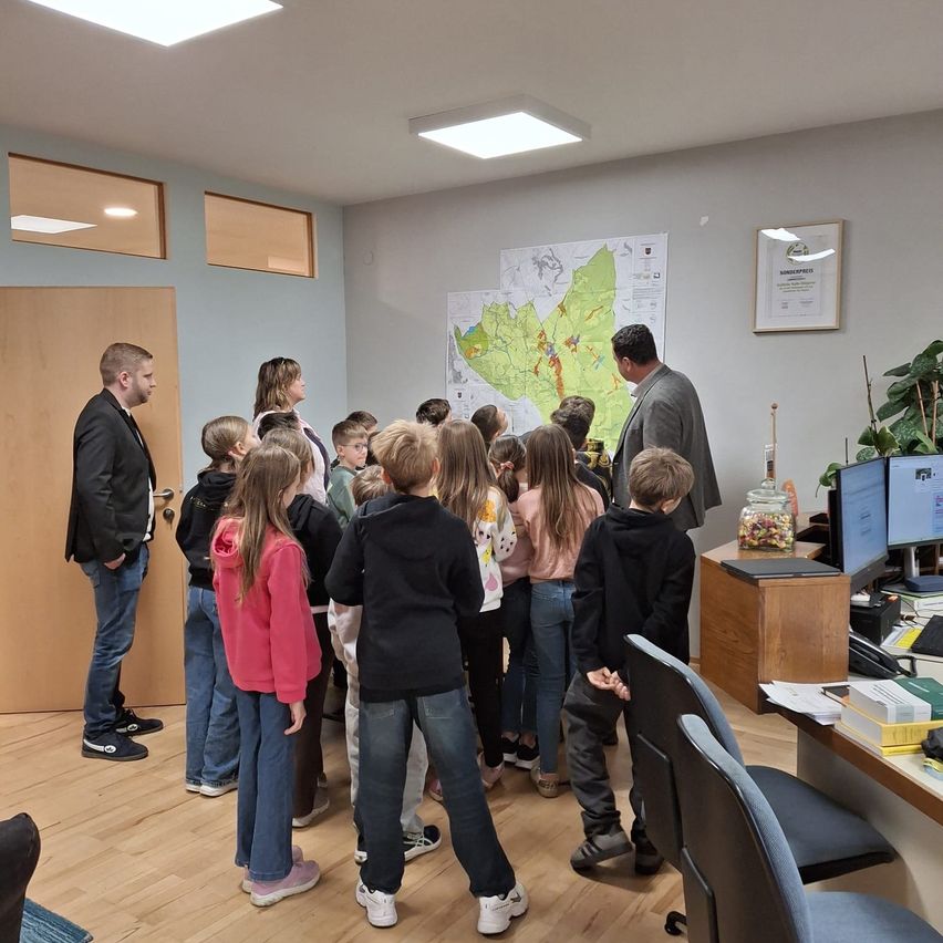 Eine Gruppe von Kindern und zwei Erwachsenen steht in einem Raum und betrachtet eine große Karte an der Wand. Der Raum hat Holzböden und einen Schreibtisch mit Computern.