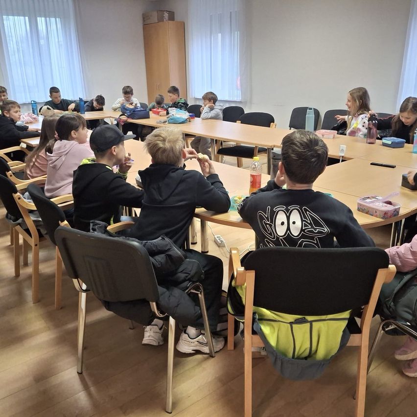 Ein Klassenzimmer mit mehreren Schülern, die an Tischen sitzen und Aktivitäten nachgehen. Die Tische sind mit Gegenständen wie Flaschen und Schachteln ausgestattet. Der Raum hat Holzböden und weiße Wände.