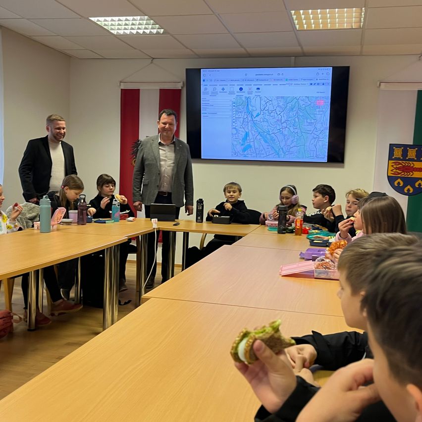 Ein Klassenzimmer mit Kindern an Tischen sitzend, die essen, zwei Männer stehen vorne neben einer großen Leinwand, die eine Karte zeigt.