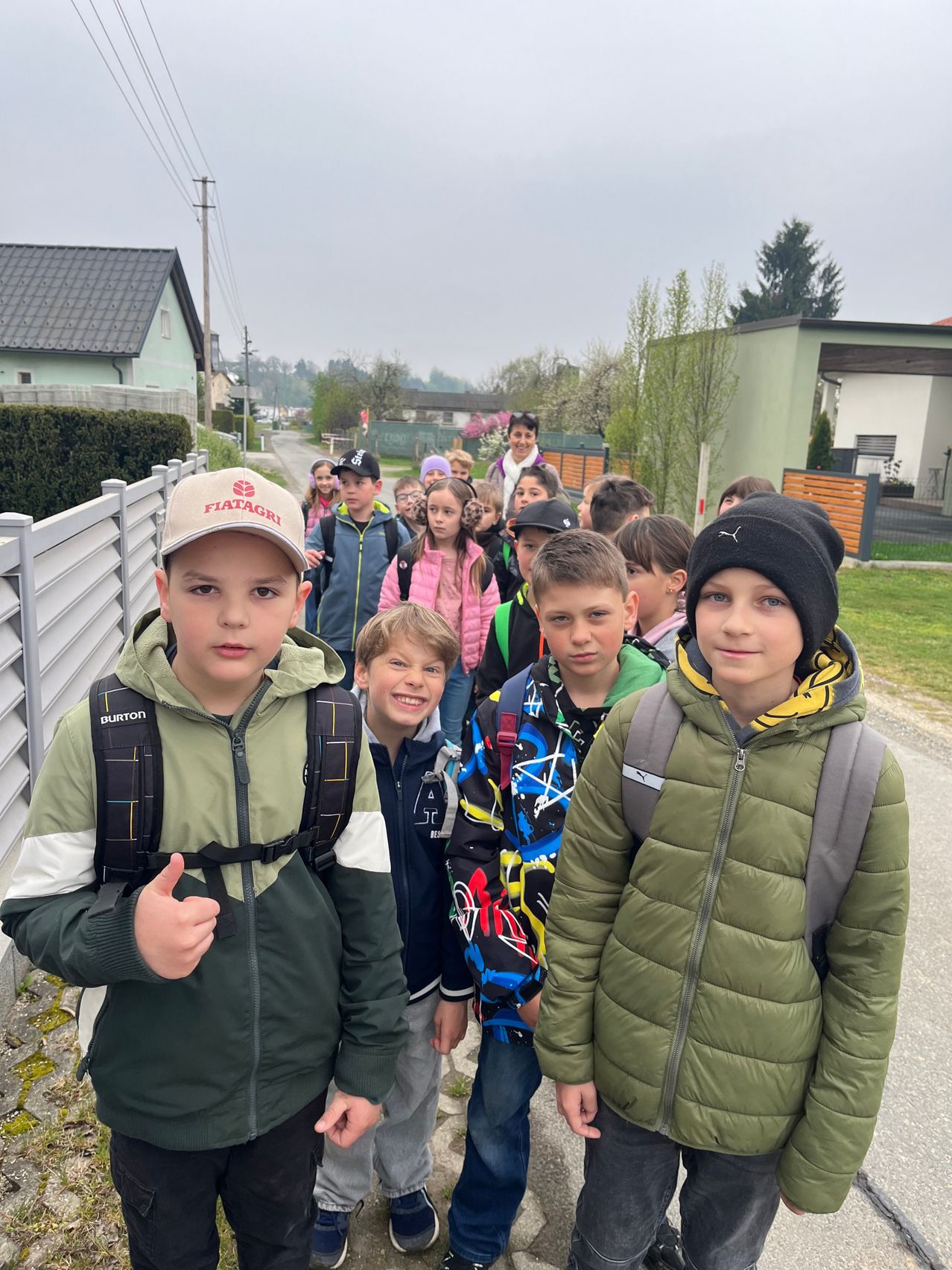 Eine Gruppe von Kindern in Winterjacken, Mützen und Rucksäcken geht auf der Straße. Sie werden von Erwachsenen begleitet, und Häuser und Bäume sind im Hintergrund.