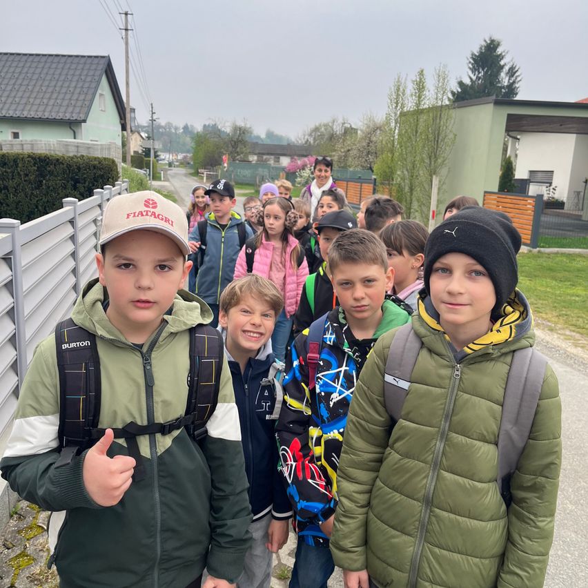 Eine Gruppe von Kindern in Winterjacken, Mützen und Rucksäcken geht auf der Straße. Sie werden von Erwachsenen begleitet, und Häuser und Bäume sind im Hintergrund.