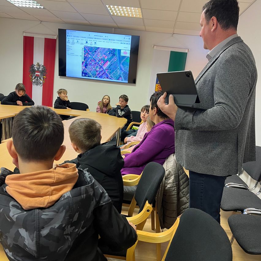 Ein Mann mit einem Tablet steht vor einer Klasse von Schülern, mit einem großen Bildschirm, der eine Karte hinter ihm anzeigt.